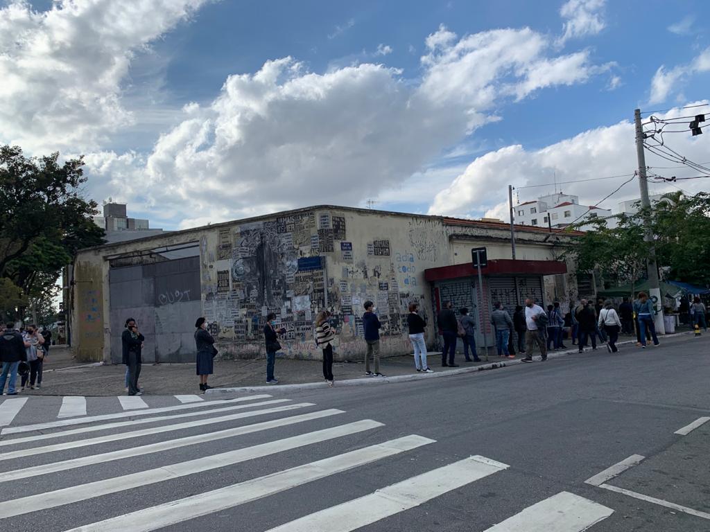 Numa calçada de esquina pessoas formam uma fila com distanciamento entre uma e outra, o céu está azul com nuvens e há um muro com cartazes apagados ao fundo